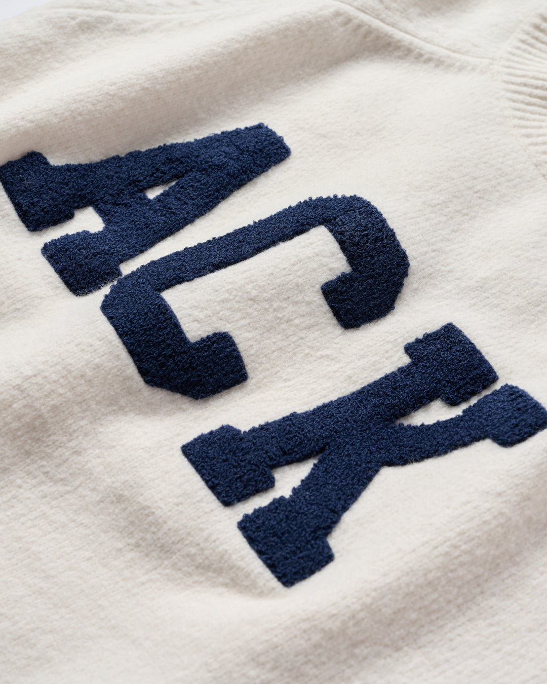 Close-up of an ivory cream-colored knit sweater featuring large, raised navy blue chenille collegiate-style letters spelling &