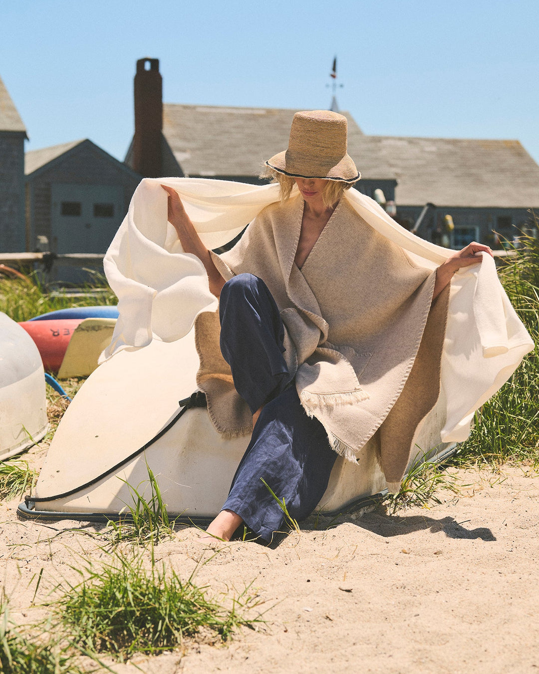 A woman sitting on sand outdoors under a straw hat, wrapped in a large cream-colored wrap and oatmeal cape, with blue pants and a beach house in the background. Amelia Cape in Oatmeal by Ellsworth + Ivey.