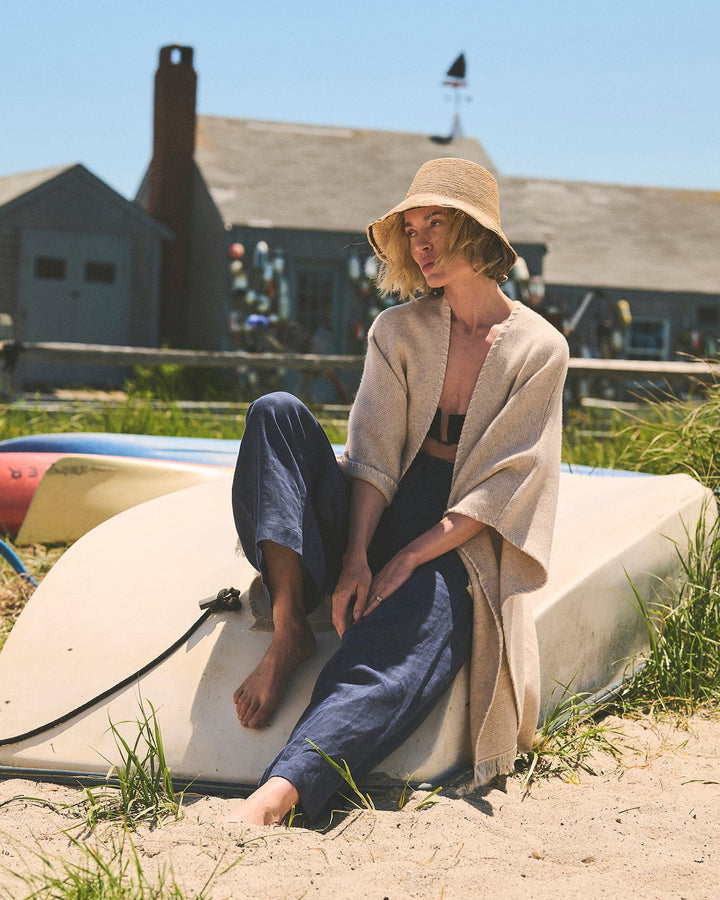 A woman sitting on sand outdoors under a straw hat, wrapped in a large cream-colored wrap and oatmeal cape, with blue pants and a beach house in the background. Amelia Cape in Oatmeal by Ellsworth + Ivey.
