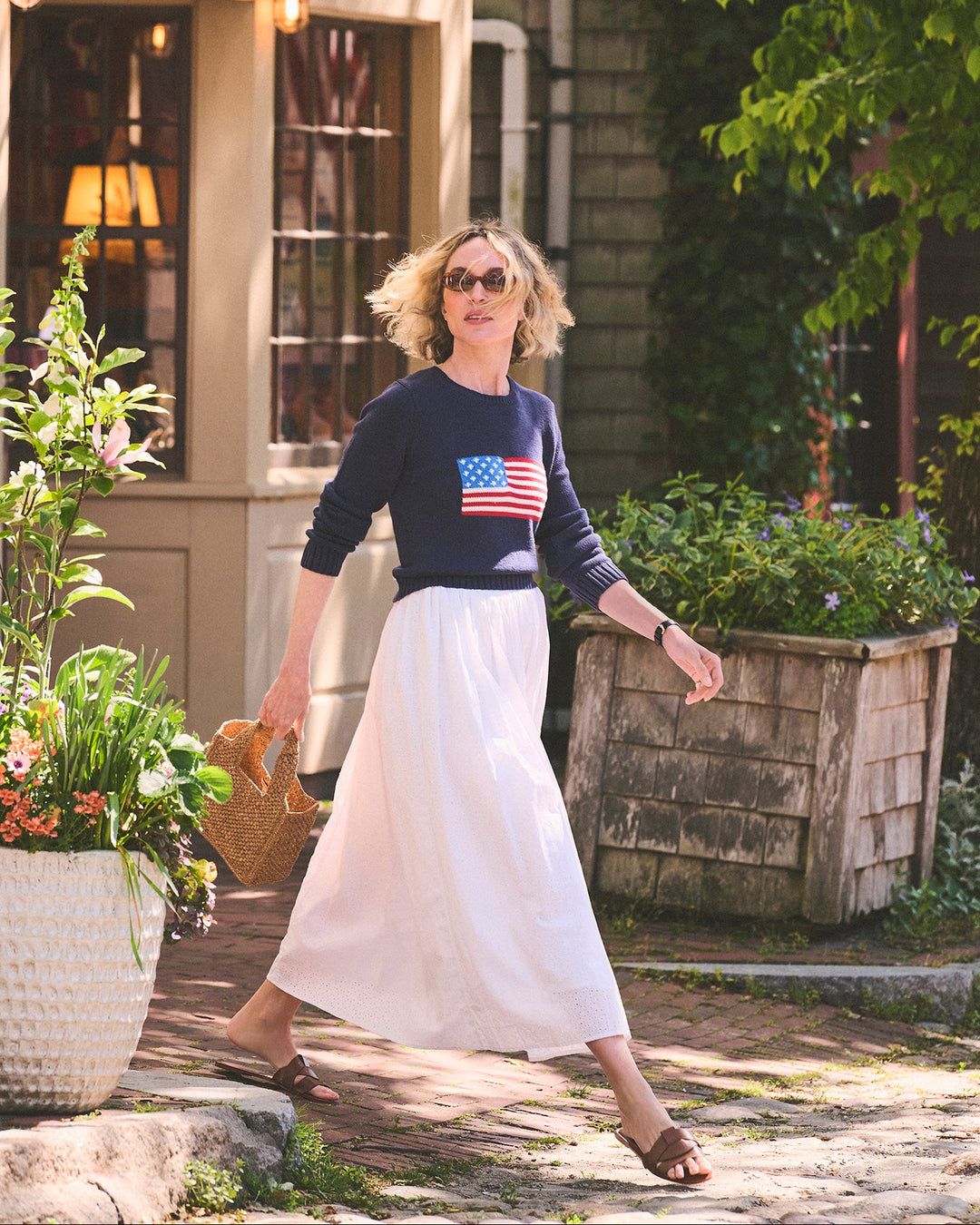 A woman walking outdoors in front of a shop in Nantucket. She is wearing a navy long-sleeve navy sweater with an American flag graphic on the chest and a long white skirt. Autumn Crew Sweater - American Flag by Ellsworth + Ivey.