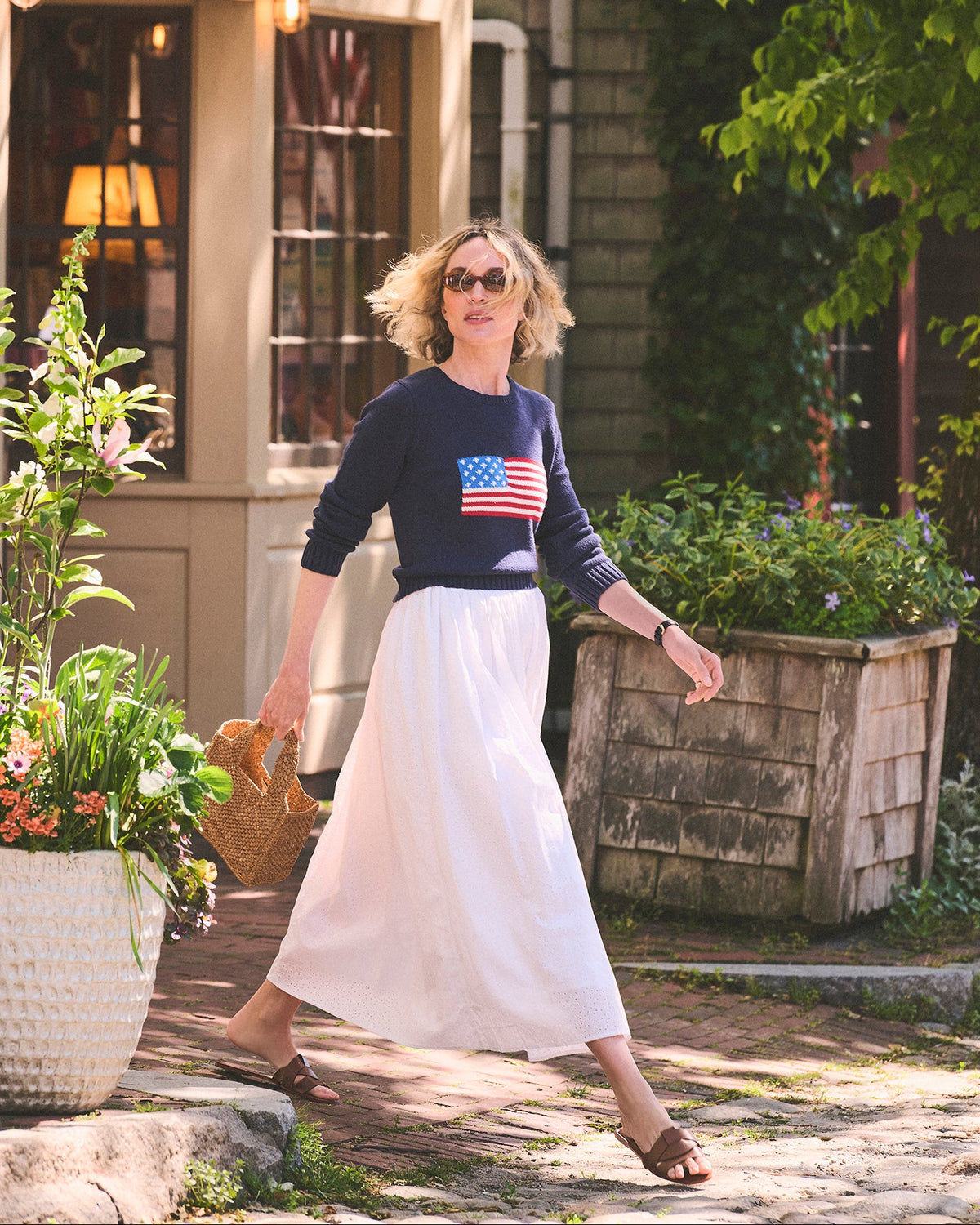 A woman walking outdoors in front of a shop in Nantucket. She is wearing a navy long-sleeve navy sweater with an American flag graphic on the chest and a long white skirt. Autumn Crew Sweater - American Flag by Ellsworth + Ivey.