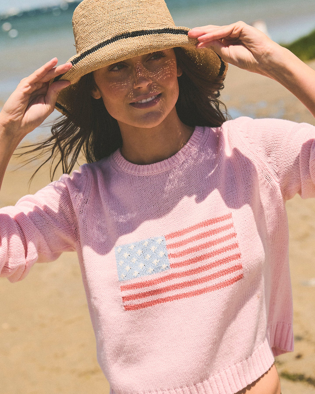 A woman on a beach wearing a light pink knit sweater with a red, white, and blue American flag graphic on the front, along with a straw hat. Autumn Crew - American Flag by Ellsworth + Ivey.