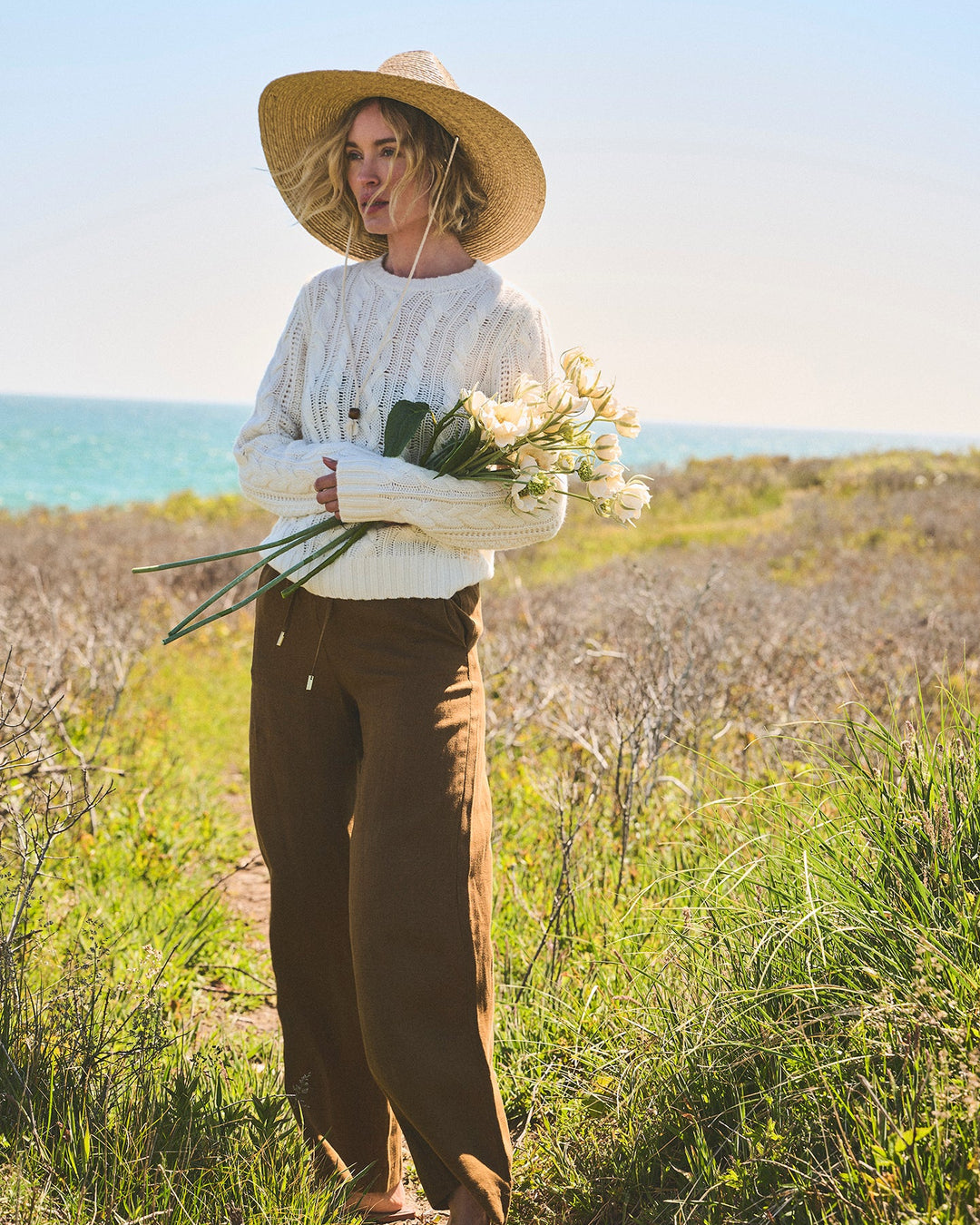 A woman stands outdoors in a grassy coastal field wearing an ivory cream cable-knit crewneck sweater, brown wide-leg pants, a straw hat, and holding a bouquet of white flowers. Carolina Cable Knit Sweater in Ivory by Ellsworth + Ivey.