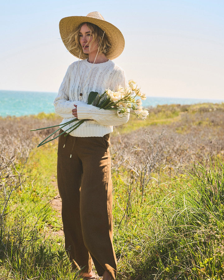 A woman stands outdoors in a grassy coastal field wearing an ivory cream cable-knit crewneck sweater, brown wide-leg pants, a straw hat, and holding a bouquet of white flowers. Carolina Cable Knit Sweater in Ivory by Ellsworth + Ivey.