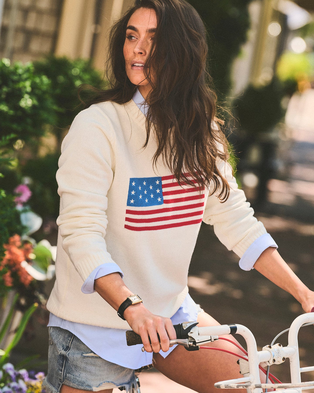 A woman riding a white bicycle outdoors wearing a cream-colored crewneck sweater with a printed American flag on the chest, layered over a light blue collared shirt and denim shorts. Classic Crew Sweater - American Flag by Ellsworth + Ivey.