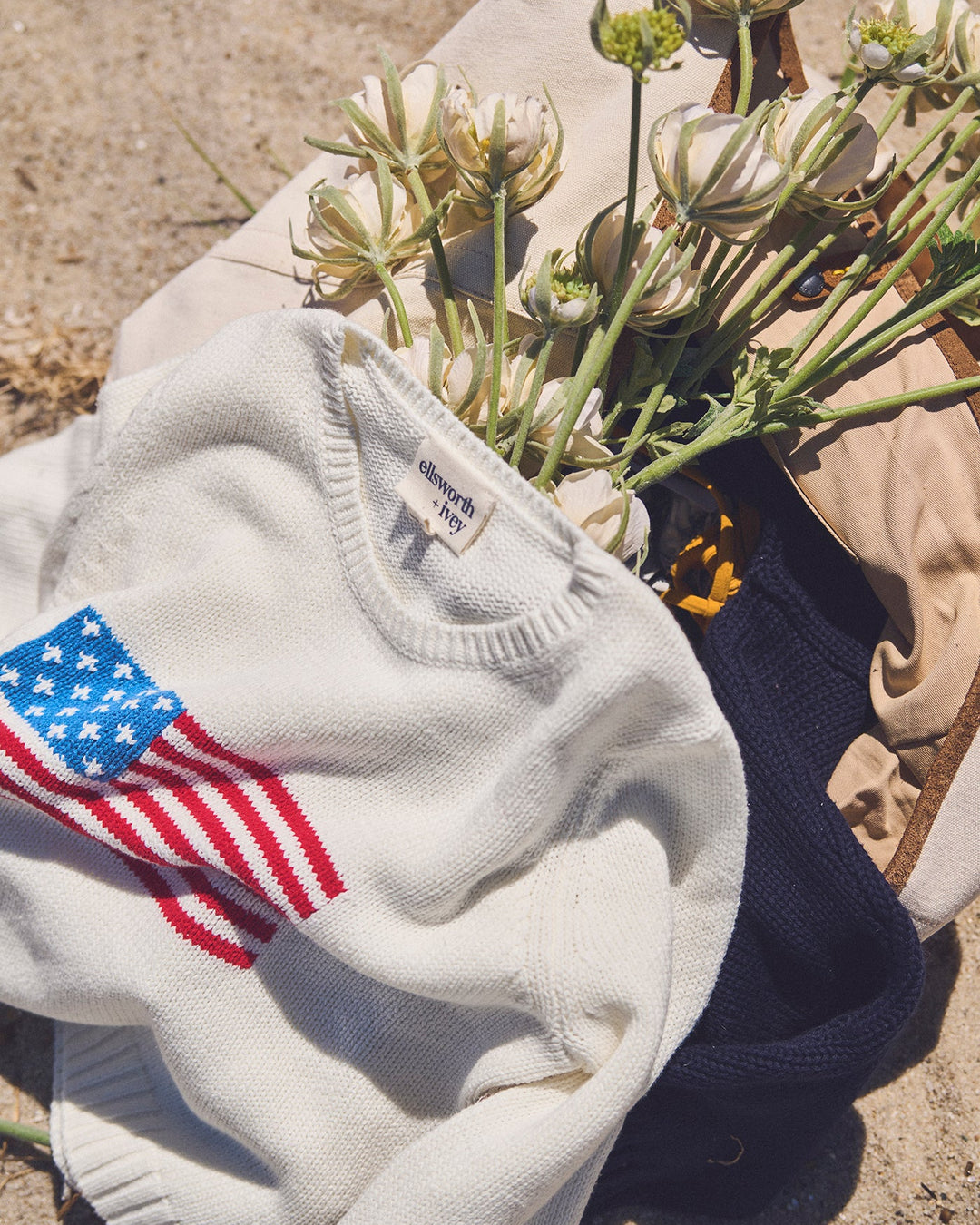 A white knit sweater with a red, white, and blue American flag graphic on the sleeve, styled outdoors with dried white flowers on a sandy surface. Classic Crew Sweater - American Flag by Ellsworth + Ivey.
