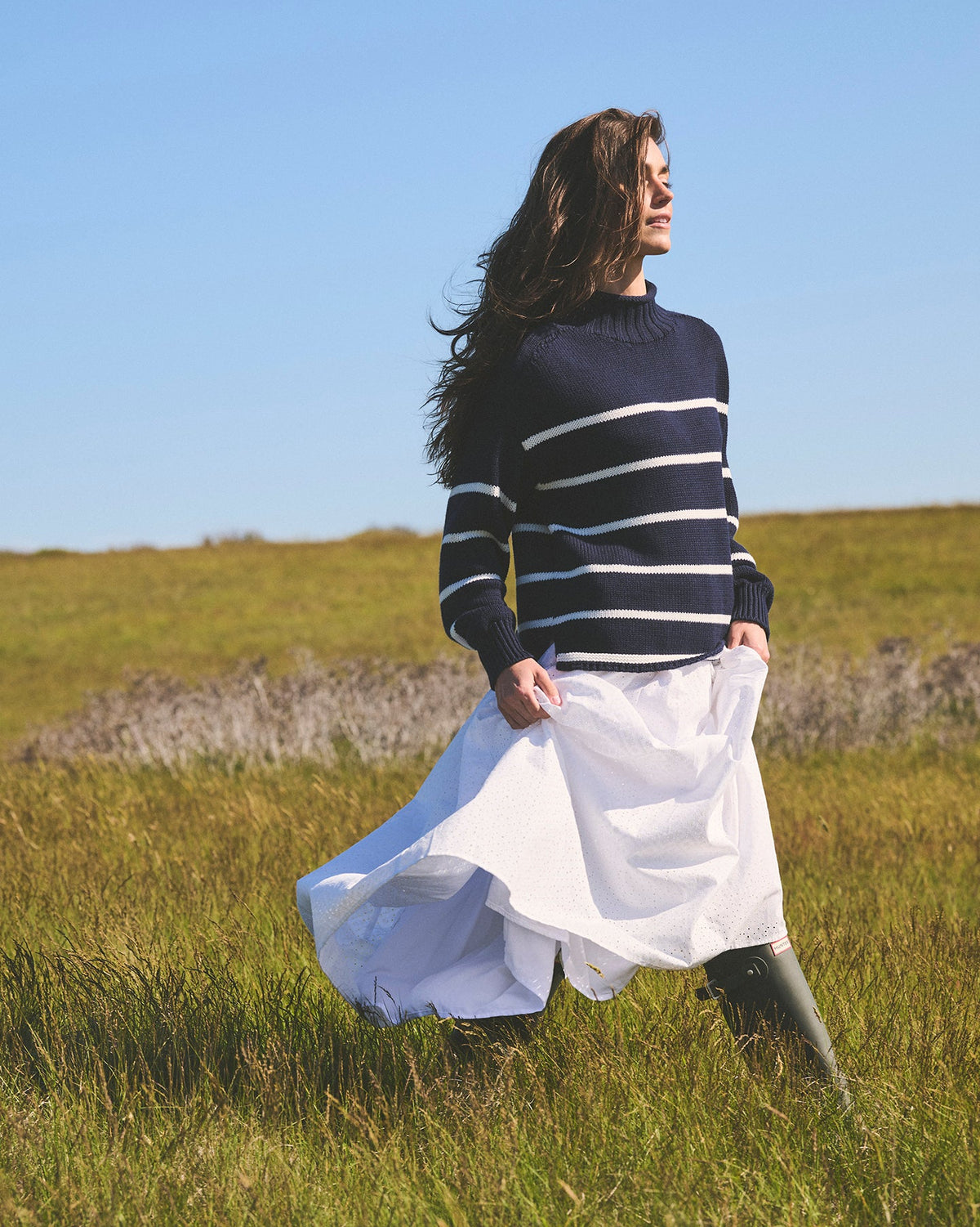 A woman stands in a grassy field under a clear blue sky, wearing a navy and white horizontally striped mock neck knit women's oversized sweater, a flowing white skirt, and dark boots. Cornelia Mock Neck Sweater in Navy by Ellsworth + Ivey.