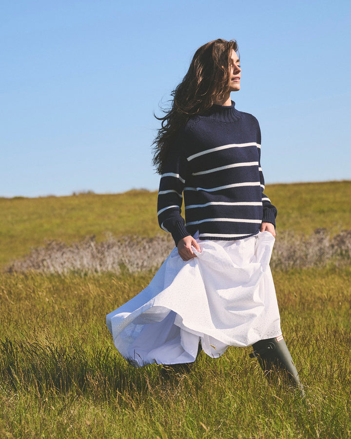 A woman stands in a grassy field under a clear blue sky, wearing a navy and white horizontally striped mock neck knit women&