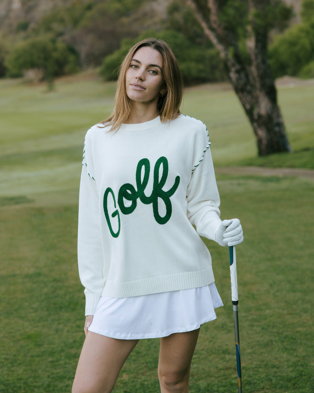 Woman in an ivory off-white knit sweater featuring the word 'Golf' embroidered in green script across the chest, with green dashed line details along the shoulders, on a golf course. Riley Crew - Golf Script by Ellsworth + Ivey.