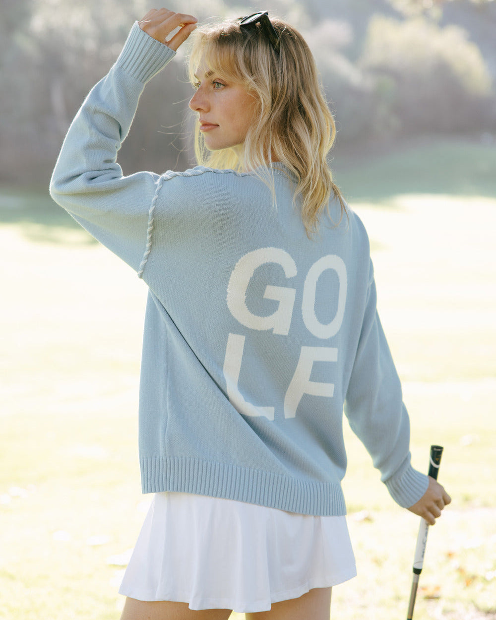 Woman in a light blue oversized crewneck sweater with 'GOLF' text, white skirt, and sunglasses on head, standing on a golf course. Riley Crew - Golf Stacked by Ellsworth + Ivey.