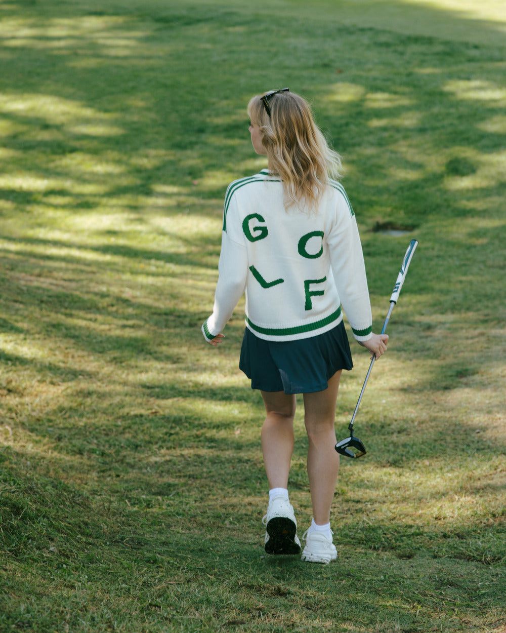 Woman in an ivory off-white golf cardigan sweater on a golf course holding a golf club. Emily Varsity Cardigan - Golf by Ellsworth + Ivey.