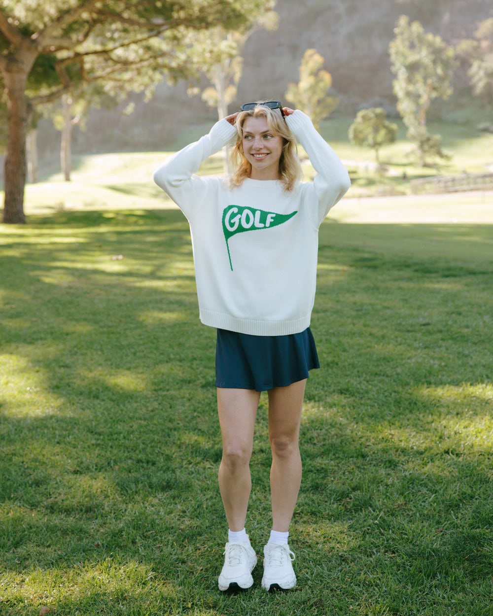 Person wearing an ivory off-white long-sleeve crewneck women's sweater featuring a green flag graphic on the chest with the word 'GOLF' in white, standing on grass with trees in the background. Riley Crew - Golf Pennant by Ellsworth + Ivey.