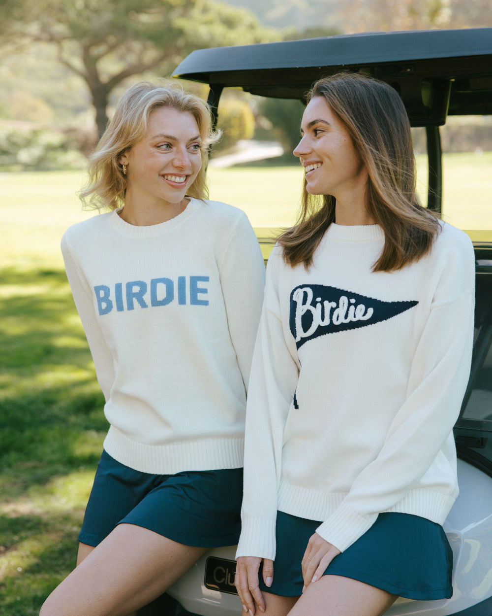 Two women wearing ivory off-white 'Birdie' sweaters sitting on a golf cart in a scenic outdoor setting.  Riley Crew - Birdie Pennant by Ellsworth + Ivey.