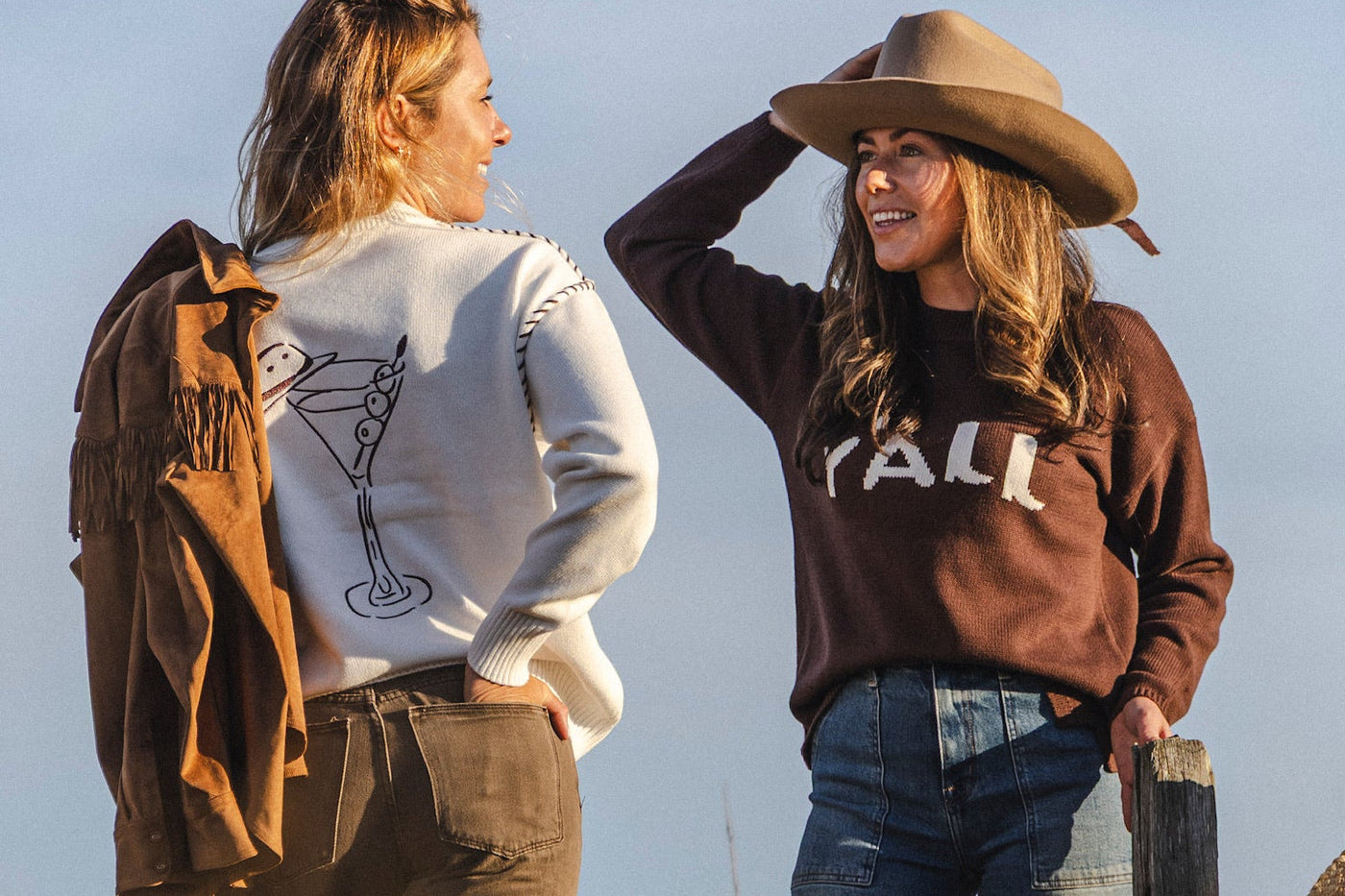 Two women in western-inspired sweaters, one in a white sweater with a martini glass design and the other in a brown “Y’all” sweater, leaning on a wooden fence in a golden field.