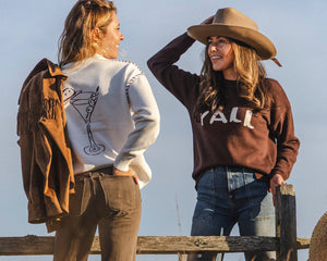 Two women in western-inspired sweaters, one in a white sweater with a martini glass design and the other in a brown “Y’all” sweater, leaning on a wooden fence in a golden field.