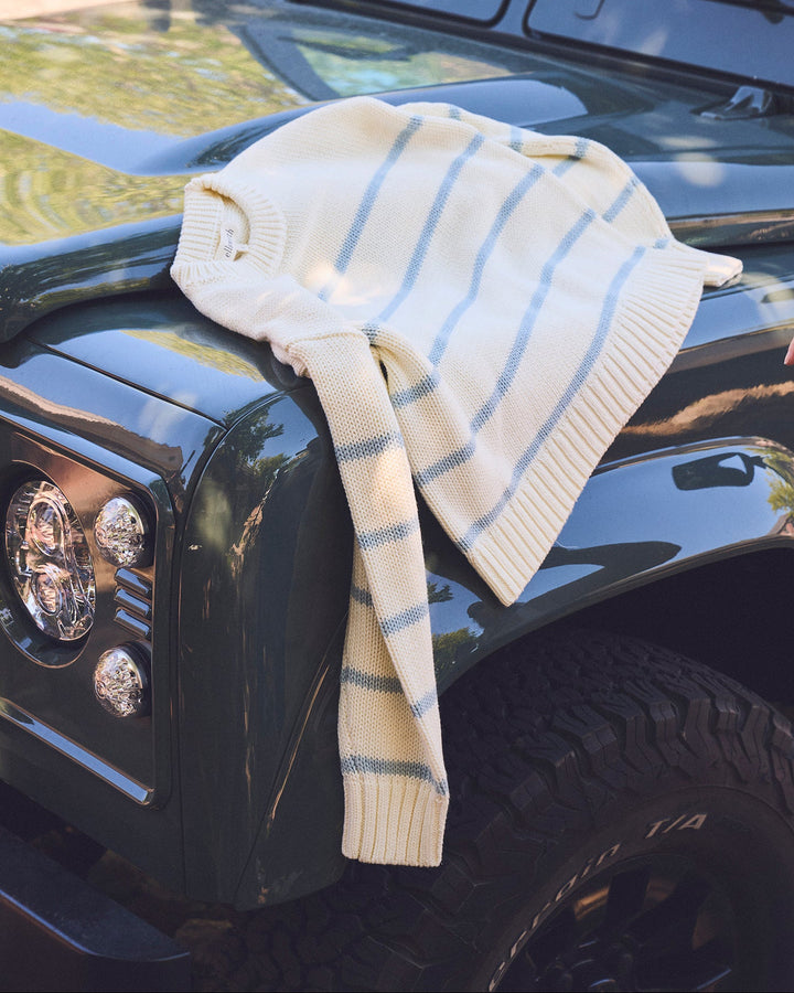 A cream-colored knit sweater with thin blue stripes draped over the hood of a dark green off-road vehicle. Elena Crew Sweater in Cornflower Blue/ Ivory by Ellsworth + Ivey.