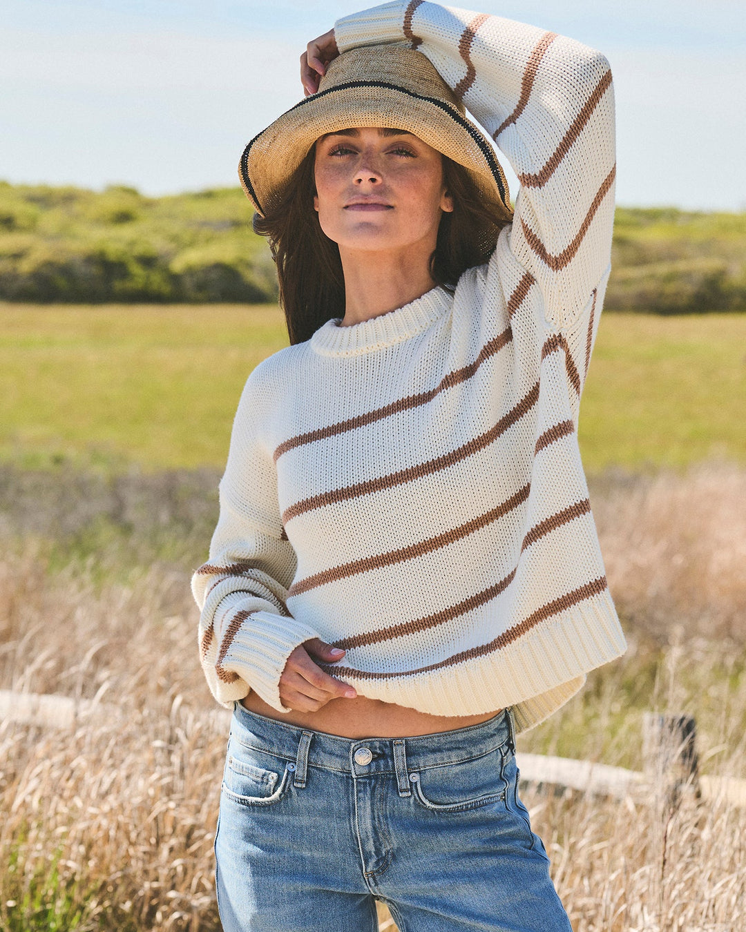 A woman wearing a cream knit sweater with brown horizontal stripes, a wide-brim straw hat, and blue jeans, standing outdoors in a grassy field. Elena Crew Sweater in Ivory/Mocha by Ellsworth + Ivey.