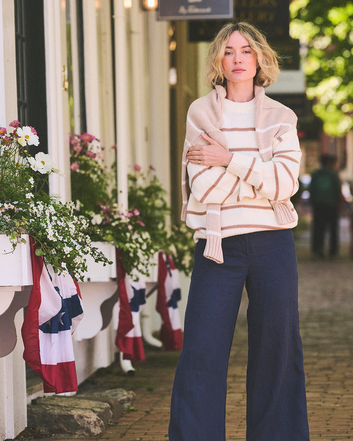 A woman stands on a brick sidewalk in front of a storefront, wearing a cream and tan striped knit cardigan over a light top, and navy wide-leg pants. Elena Crew Sweater in Ivory/Mocha by Ellsworth + Ivey.