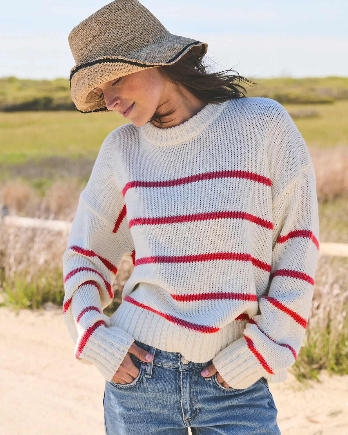 A woman wearing an ivory cream knit crewneck sweater with red horizontal stripes, blue jeans, and a straw hat, standing outdoors with grassy dunes in the background. Elena Crew Sweater in Ivory/Red by Ellsworth + Ivey.