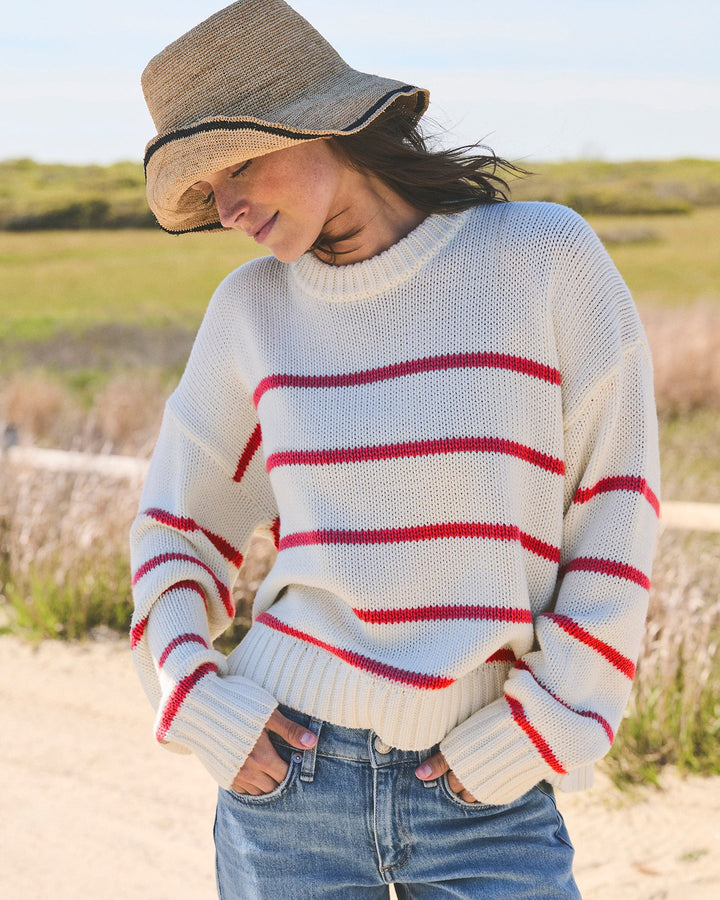 A woman wearing an ivory cream knit crewneck sweater with red horizontal stripes, blue jeans, and a straw hat, standing outdoors with grassy dunes in the background. Elena Crew Sweater in Ivory/Red by Ellsworth + Ivey.