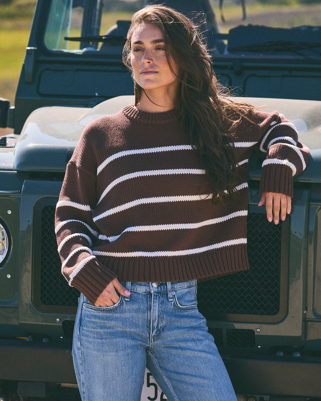 A woman wearing a warm brown and white striped crewneck knit sweater and light blue jeans, posing in front of a dark green off-road vehicle outdoors.  Elena Crew Sweater in Warm Brown / Ivory by Ellsworth + Ivey. 
