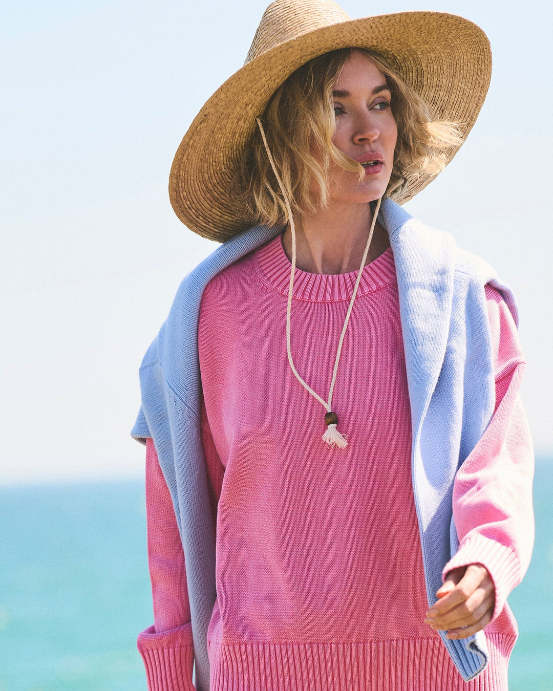 A woman wearing a pink crewneck sweater, a light blue cardigan, a wide-brimmed straw hat, and a shell pendant necklace, standing outdoors with the ocean in the background. Emery Crewneck Sweater in Bright Pink by Ellsworth &amp; Ivey.