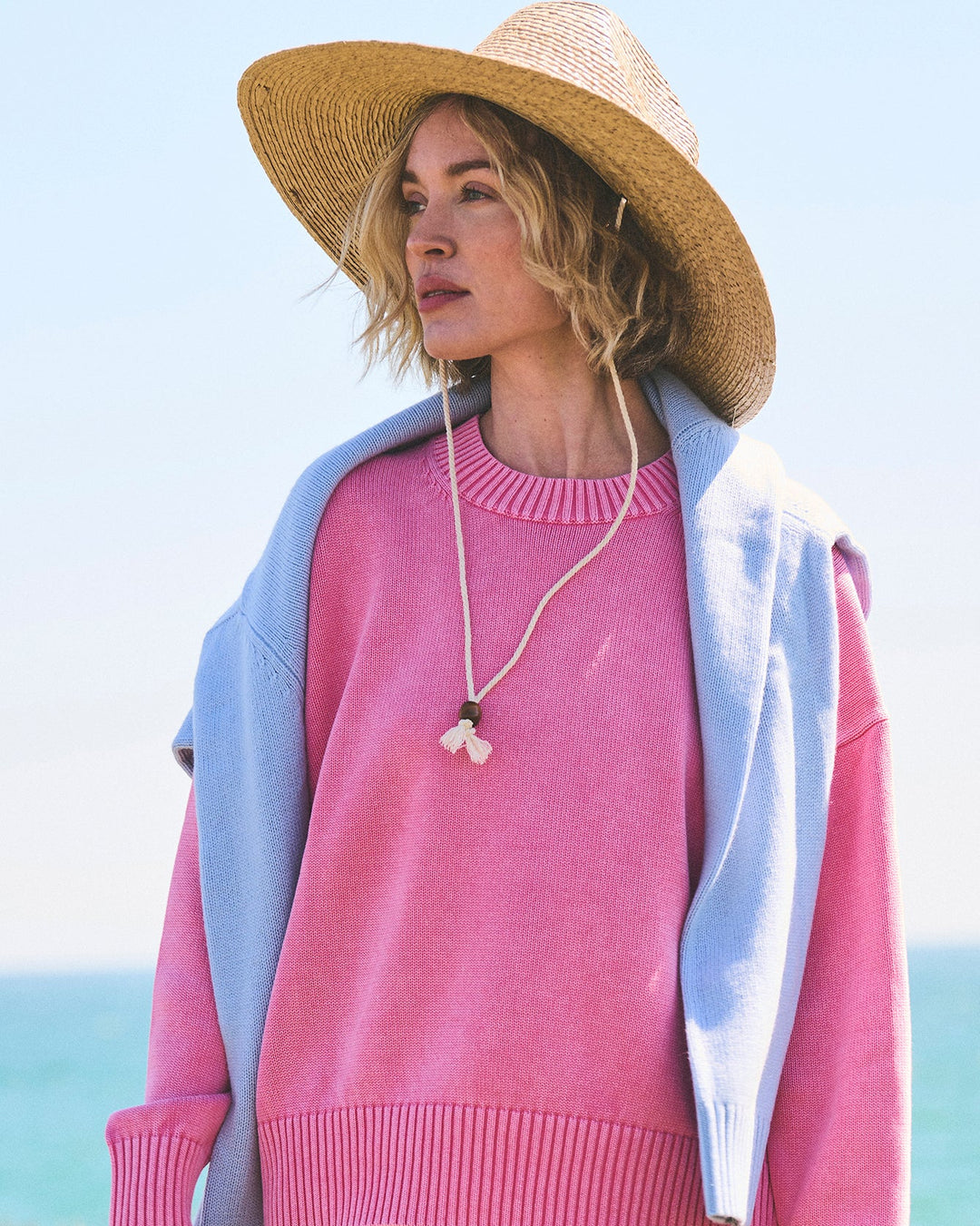 A woman standing outdoors near the beach wearing a pink ribbed knit sweater, a light blue cardigan, a wide-brim straw hat, and cream bikini bottoms. Emery Crewneck Sweater in Bright Pink by Ellsworth &amp; Ivey.