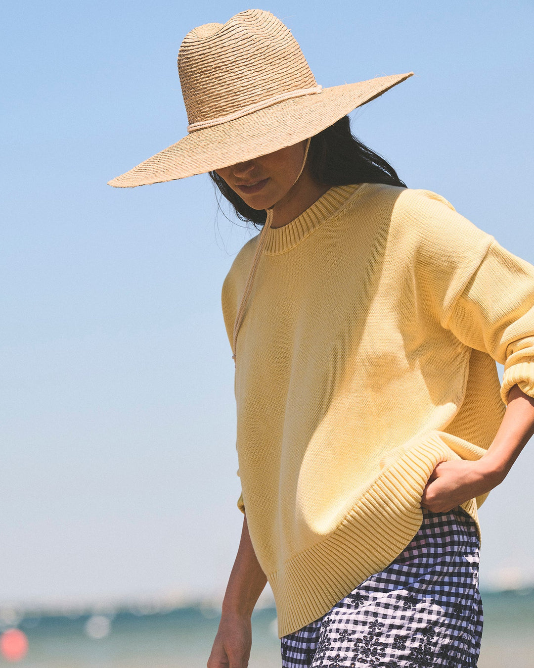 A woman wearing a wide-brimmed straw hat, a pale yellow knit sweater, and a navy and white gingham skirt, photographed outdoors against a soft-focus beach and sky background. Emery Crewneck Sweater in Butter Yellow by Ellsworth &amp; Ivey.