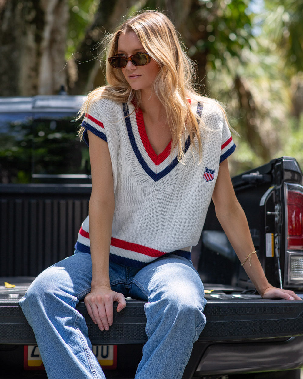 Woman wearing a white Americana sweater vest with red and blue accents and sunglasses, standing outdoors. McKenna Sweater Vest - American Shield by Ellsworth+ Ivey.