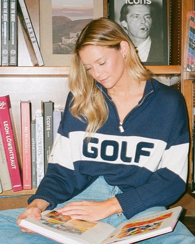 Woman in a navy blue and white colorblock mock neck sweater, reading a magazine in front of a bookshelf with 'icons' magazine. Madison Quarter Zip - Golf Colorblock by Ellsworth + Ivey.