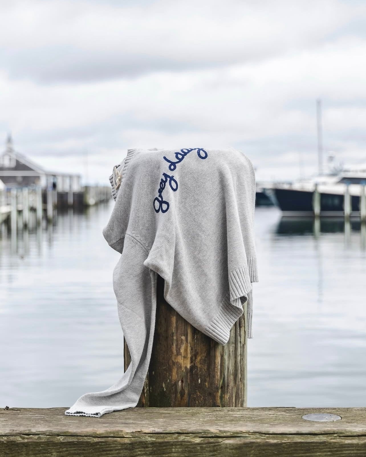 A light heather gray knit sweater with Grey Lady embroidered in navy blue script across the chest, draped over a wooden dock post. Classic Crew Sweater- Grey Lady by Ellsworth + Ivey.