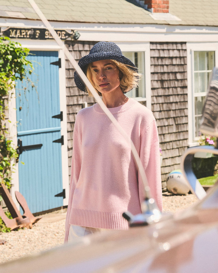 A woman wearing a light pink knit sweater and a dark blue woven hat stands outdoors in front of a shingled house with a blue door. A car hood is visible in the foreground. Joey Crew in Pastel Pink by Ellsworth + Ivey.