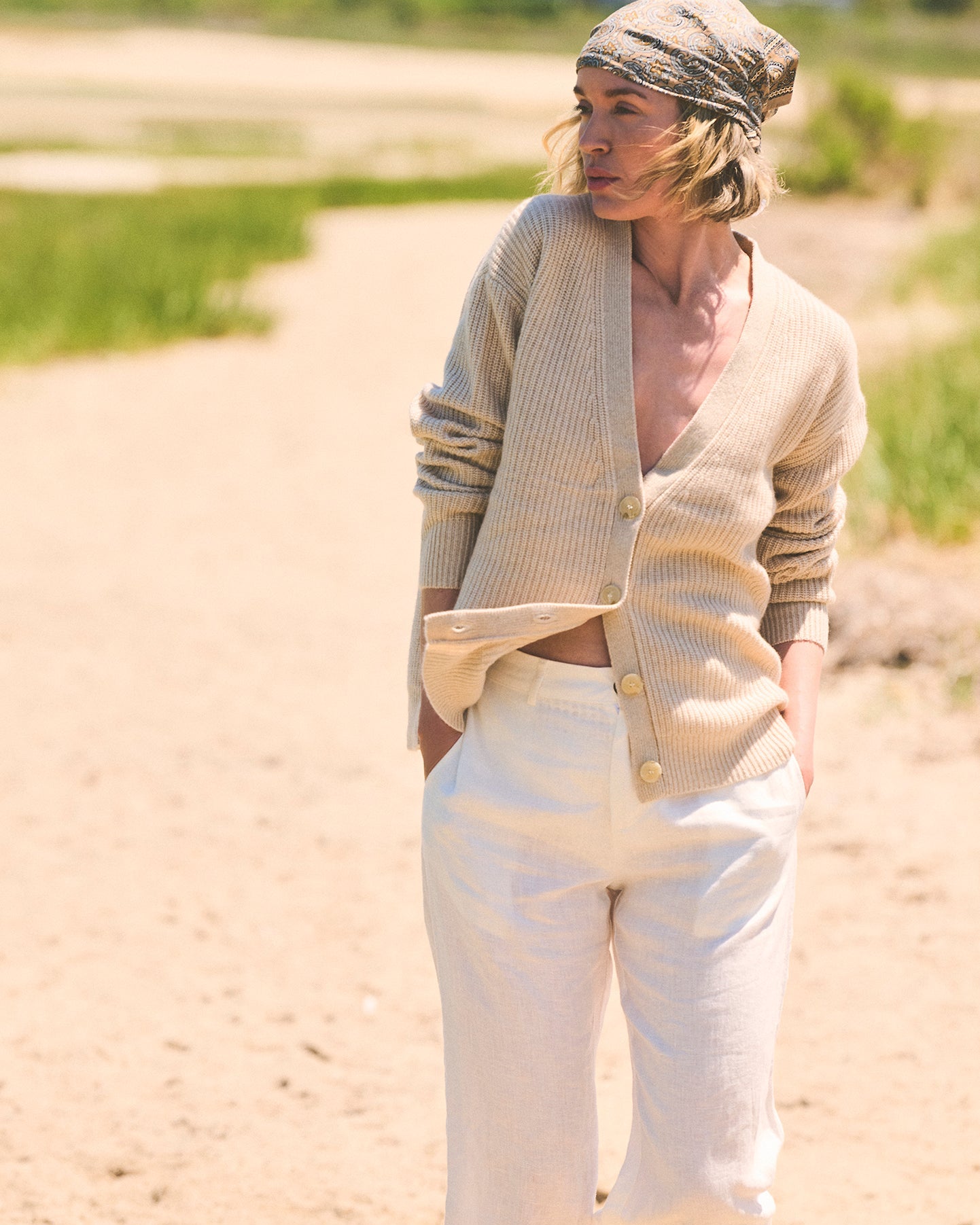 A woman standing outdoors on a sandy path wearing an oatmeal beige ribbed v-neck cashmere cardigan, white wide-leg pants, and a beige headscarf. Lilah V-Neck Knit Cardigan in Oatmeal by Ellsworth + Ivey.