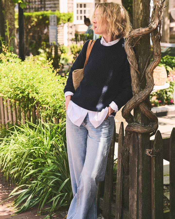 A woman stands outdoors leaning against a wooden post, wearing a dark navy sweater over a white collared shirt, light blue wide-leg jeans, and brown sandals. She has a woven shoulder bag. Quinn Fisherman Sweater in Navy by Ellsworth + Ivey.