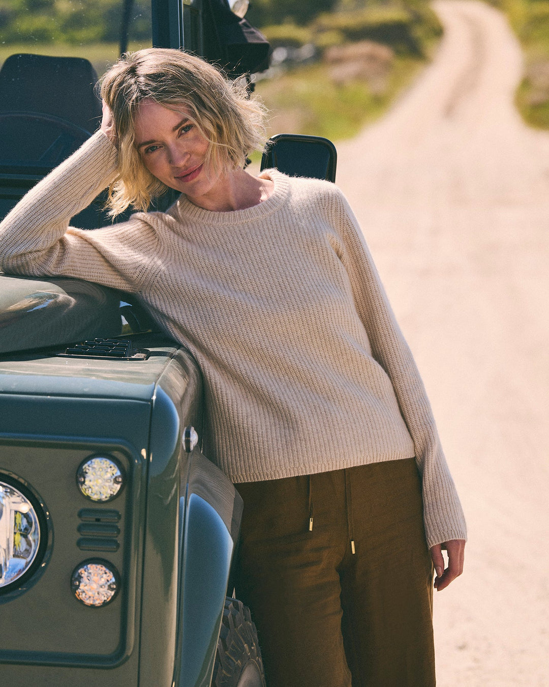 A woman with short blonde hair leans against the front of a dark green off-road vehicle on a dirt road. She is wearing an oatmeal beige ribbed knit crewneck women&