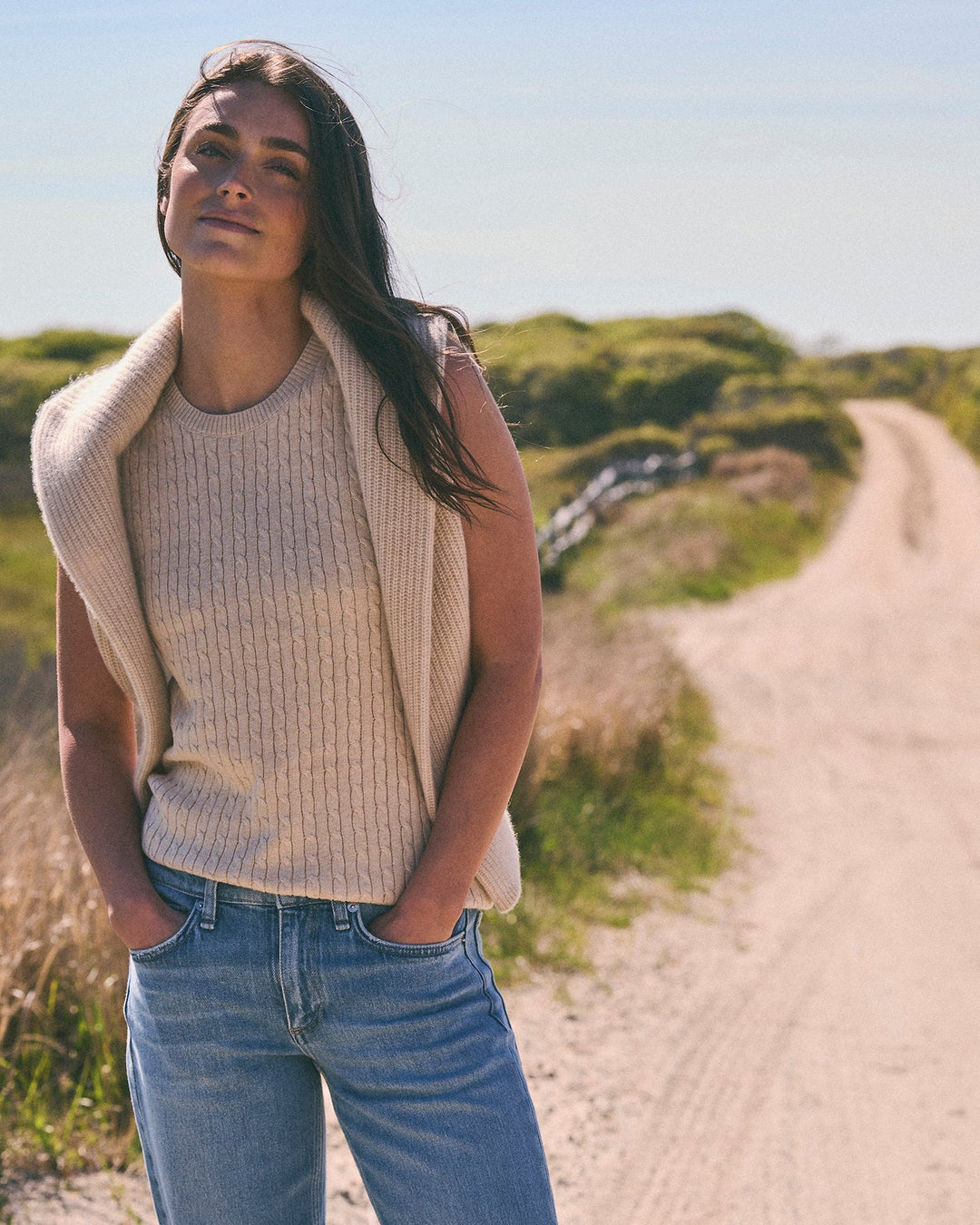 A woman standing outdoors on a sandy path wearing a sleeveless ribbed knit oatmeal tan sweater vest and blue jeans. Violet Cashmere Cable Knit Sleeveless Sweater in Oatmeal by Ellsworth + Ivey.