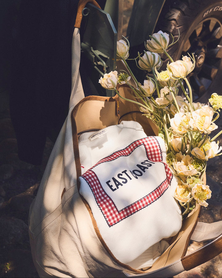 A cream canvas tote bag with tan leather handles, filled with white and cream flowers and an ivory white sweater featuring a red gingham patch that reads &