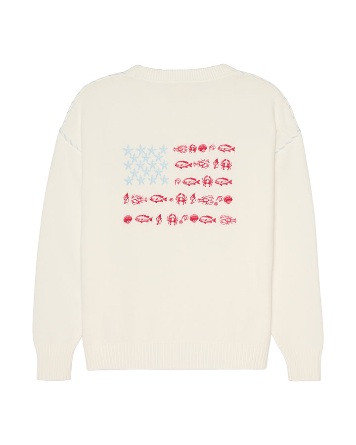 An ivory crewneck sweater shown from the back, featuring a graphic print of the American flag made from fish silhouettes in red and light blue. Ellsworth + Ivey Riley Stars and Shells Flag Sweater.