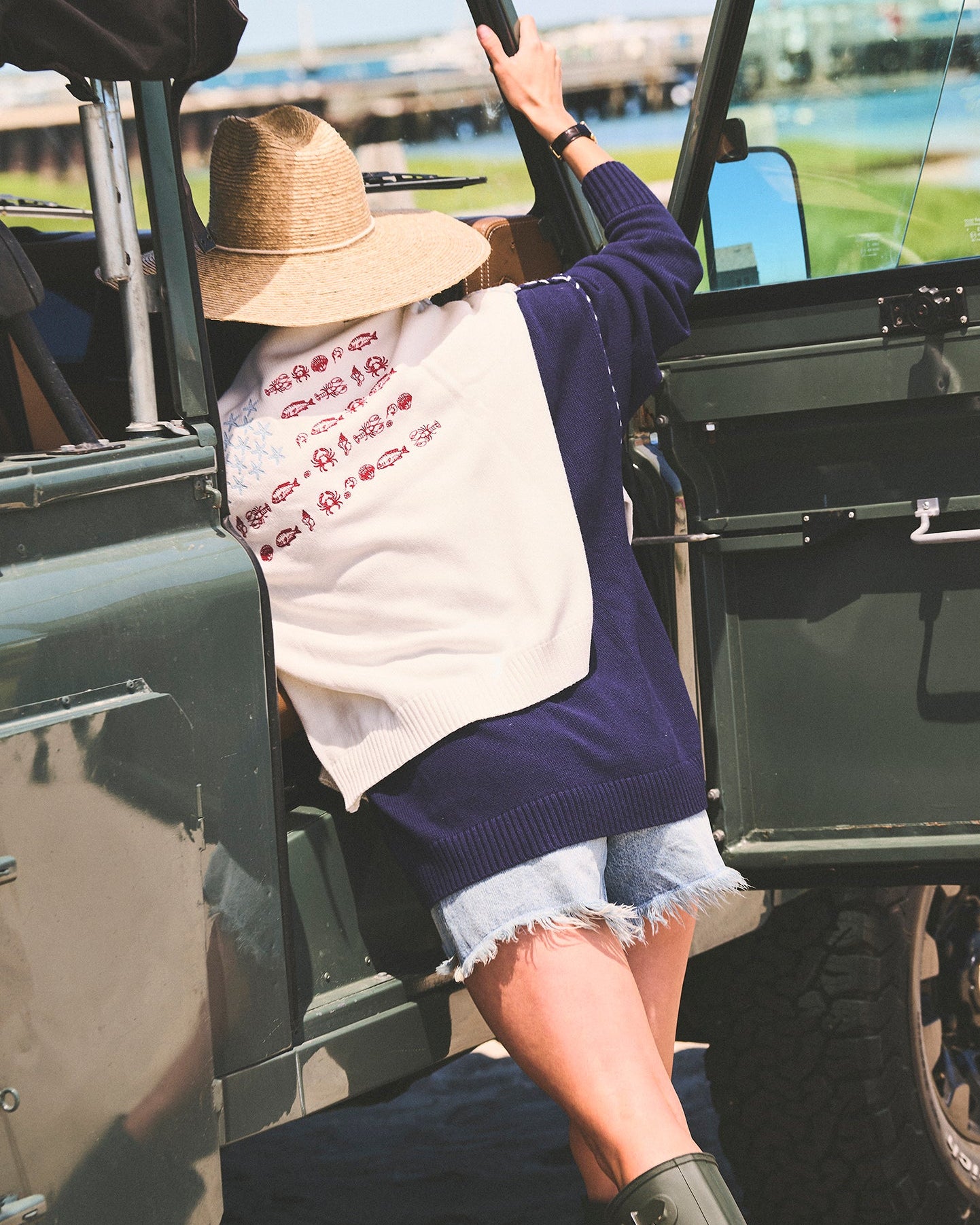A woman wearing a navy blue knit sweater with an ivory white sweater over her shoulders with stars and fish in an american flag formation, denim shorts, a straw hat, and green rain boots climbing into an open military-style green vehicle outdoors. Ellsworth + Ivey Riley Stars and Shells Flag Sweater.