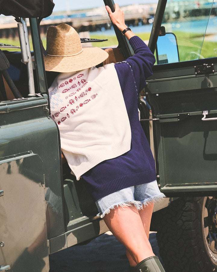 A woman wearing a navy blue knit sweater with an ivory white sweater over her shoulders with stars and fish in an american flag formation, denim shorts, a straw hat, and green rain boots climbing into an open military-style green vehicle outdoors. Ellsworth + Ivey Riley Stars and Shells Flag Sweater.