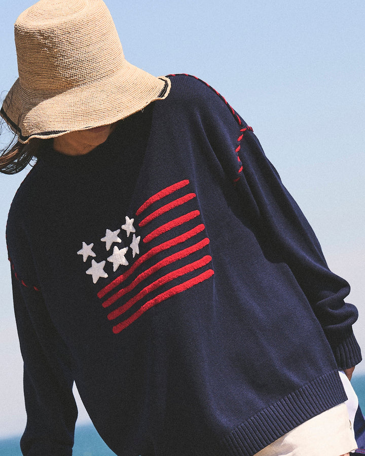 A woman wearing an oversized navy knit sweater with a red and white American flag chenille motif on the chest, paired with a straw hat and light shorts, photographed outdoors against a pale sky and water background. Riley Crew - American Flag by Ellsworth + Ivey.