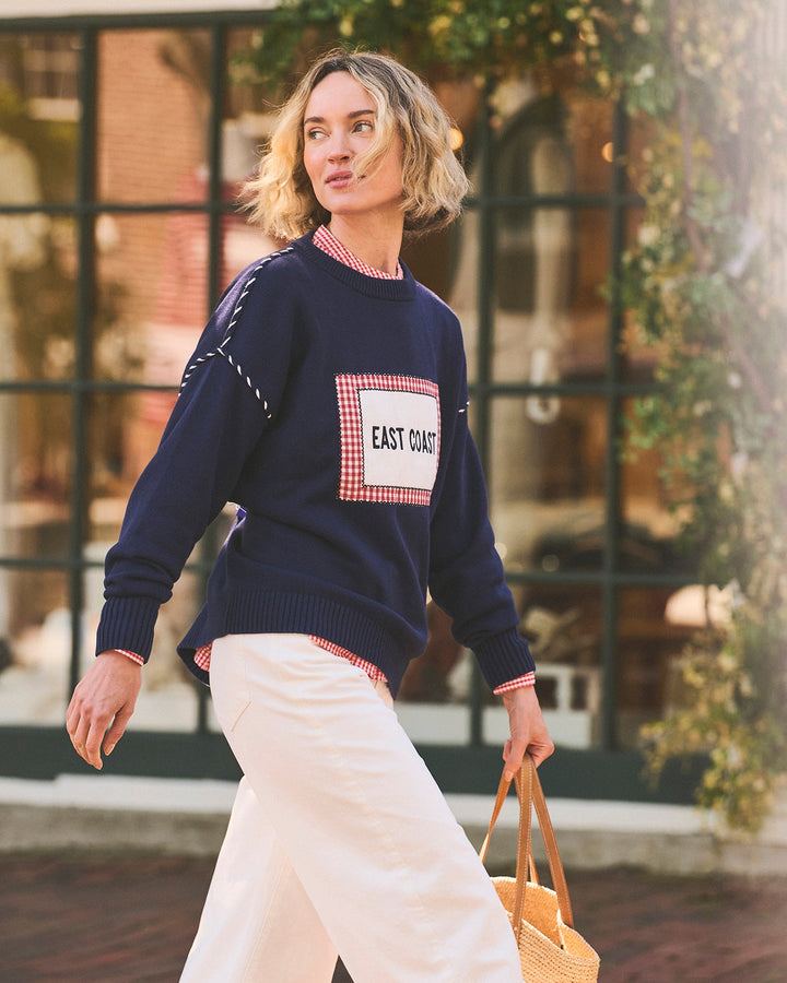 A woman walking outdoors wearing an oversized navy blue crewneck sweater with a red gingham patch on the chest that reads &