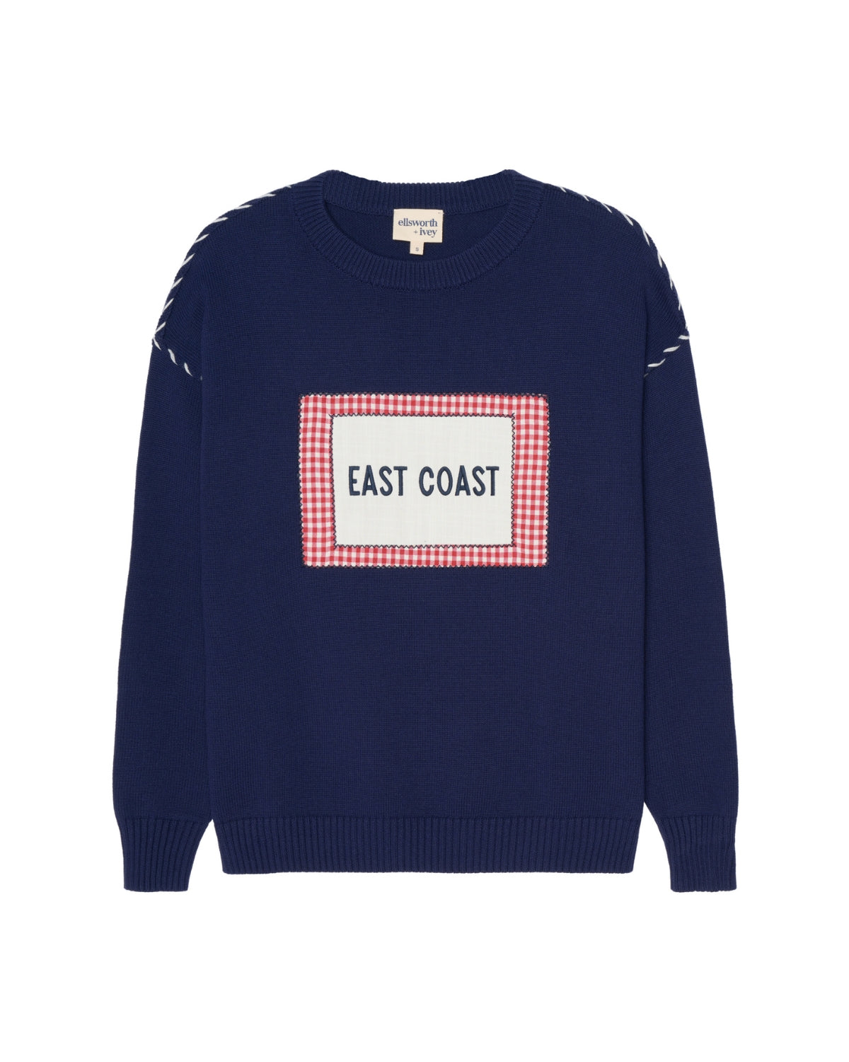 A navy blue crewneck women's sweater featuring a central rectangular patch with a red and white gingham border and the words 'EAST COAST' in navy. The sleeves have white whip stitch details near the shoulders. Ellsworth + Ivey Riley Crew - East Coast.