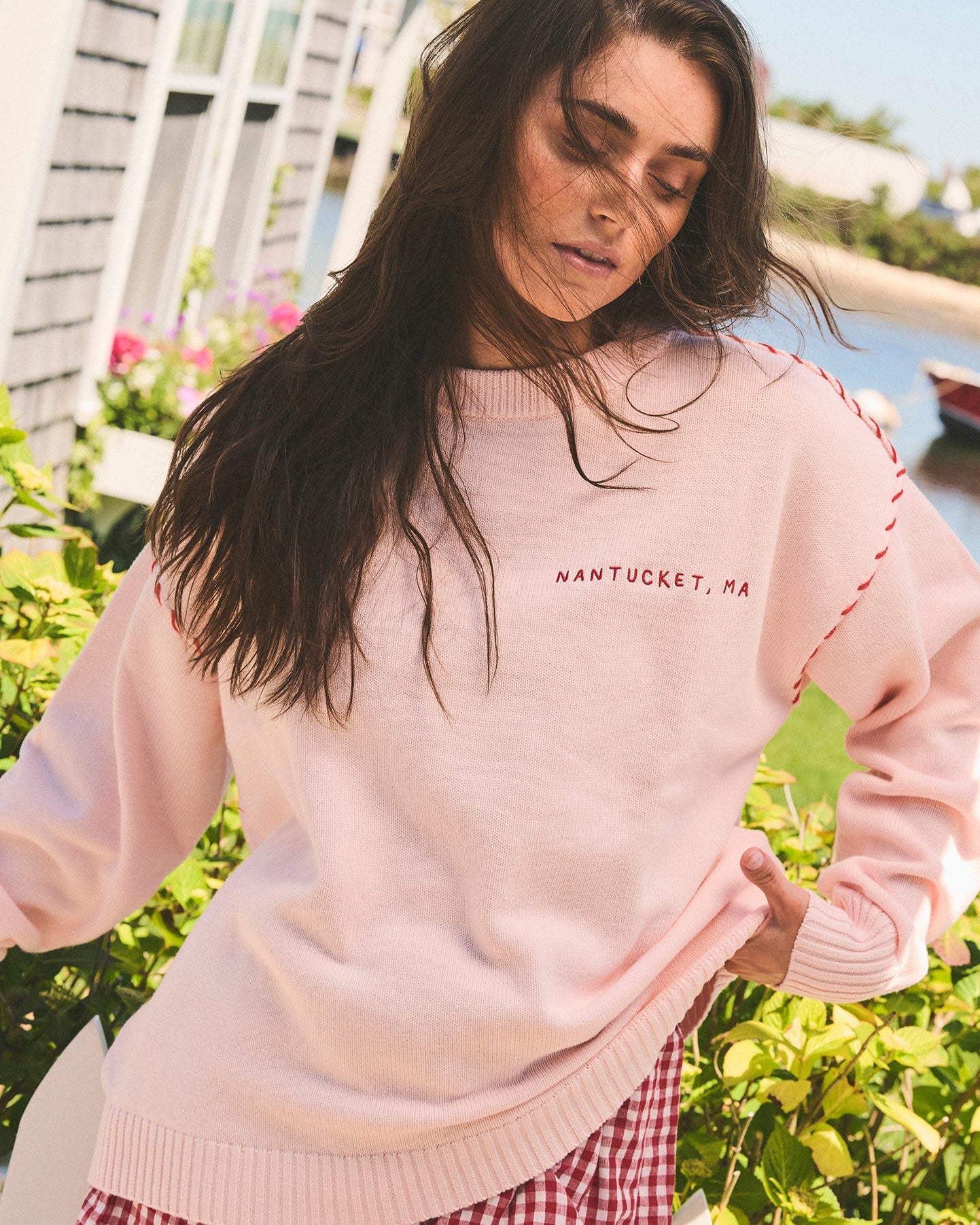 A woman wearing a light pink oversized crewneck sweatshirt with 'NANTUCKET, MA' embroidered in red on the chest, set against an outdoor background with green foliage and water. Riley Crew Sweater - ACK Lobster - Ellsworth + Ivey.