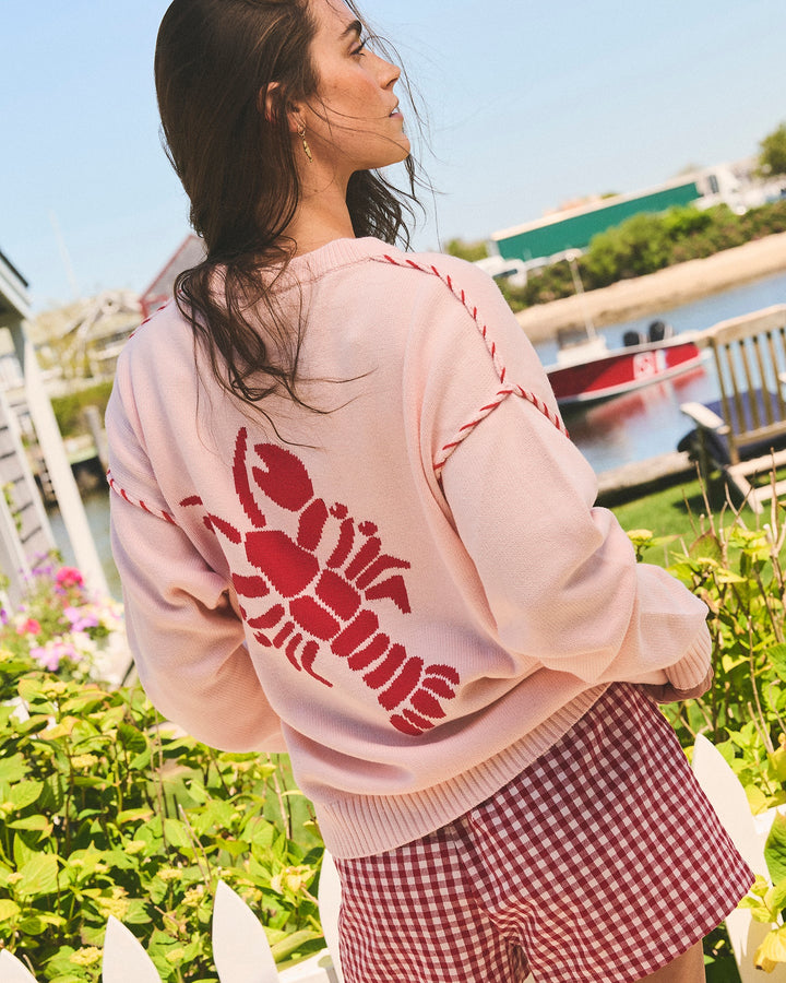 A woman wearing a light pink oversized crewneck sweatshirt with a large red lobster graphic on the back, paired with red and white gingham shorts, photographed outdoors near a white picket fence and water. Riley Crew Sweater - ACK Lobster - Ellsworth + Ivey.