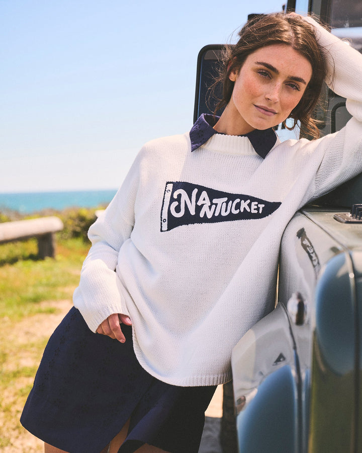 A woman wearing an  ivory white knit crewneck sweater with a blue pennant flag with Nantucket lettering, outside, leaning against a green vehicle. Sadie Crew Sweater Nantucket Pennant by Ellsworth + Ivey