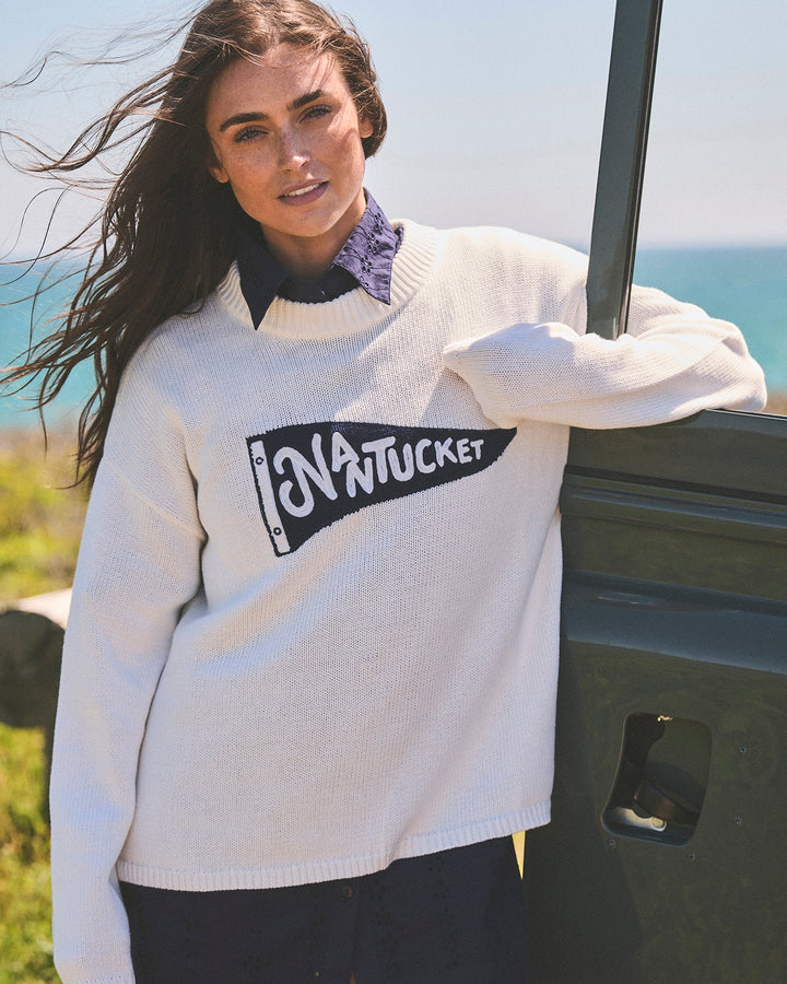 A woman wearing an  ivory white knit crewneck sweater with a blue pennant flag with Nantucket lettering, outside, leaning against a green vehicle. Sadie Crew Sweater Nantucket Pennant by Ellsworth + Ivey