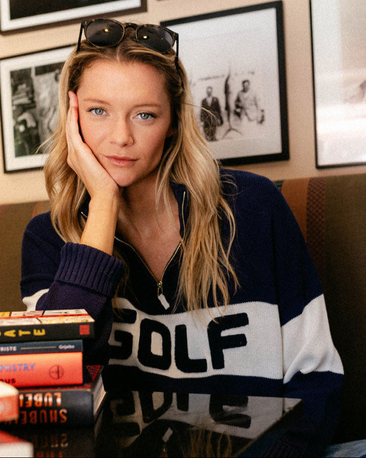 Woman wearing a navy blue and white colorblock 'GOLF' sweater with books stacked in front of her, framed pictures on the wall. Madison Quarter Zip - Golf Colorblock by Ellsworth + Ivey.