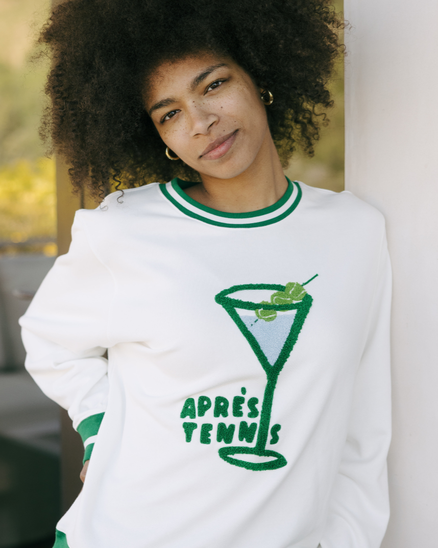 Woman wearing a white sweatshirt with a green tennis graphic and text "Apres Tennis", standing against a blurred outdoor background. Elle Apres Tennis Sweatshirt by Ellsworth + Ivey.