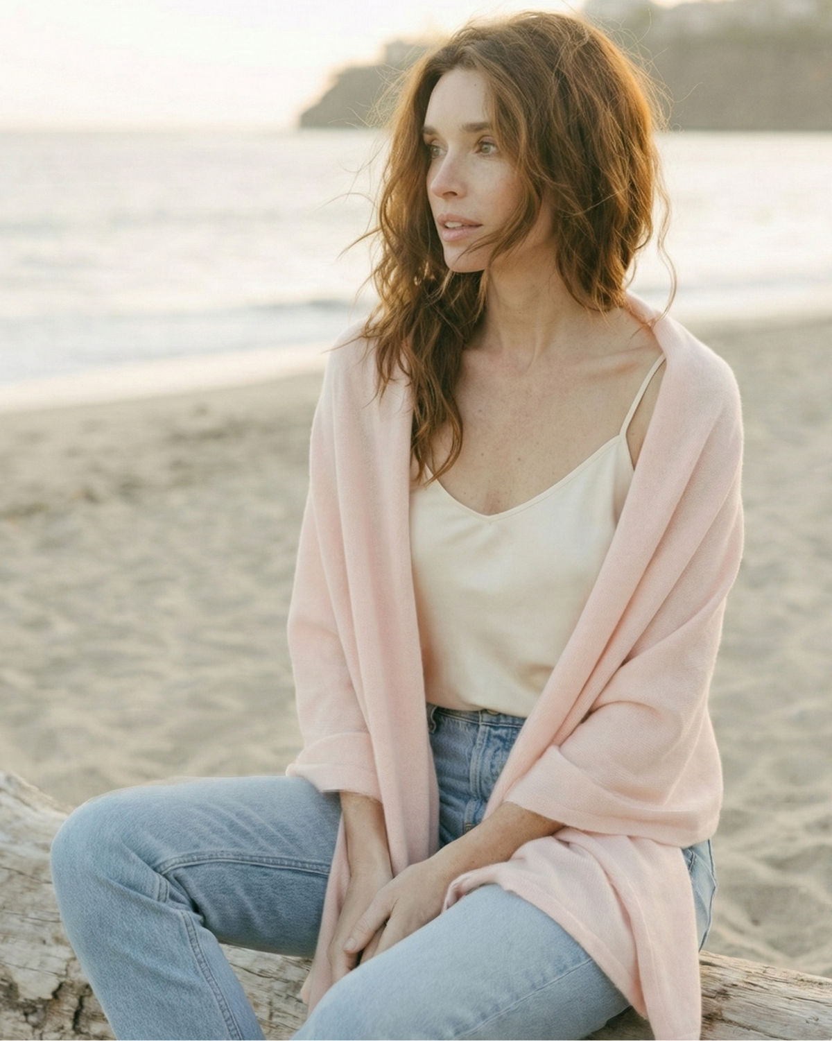 Woman wearing a white top and 
ultra soft pink wool/cashmere blend wrap scarf on a beach. Cashmere Travel Wrap in Pink by Ellsworth + Ivey.